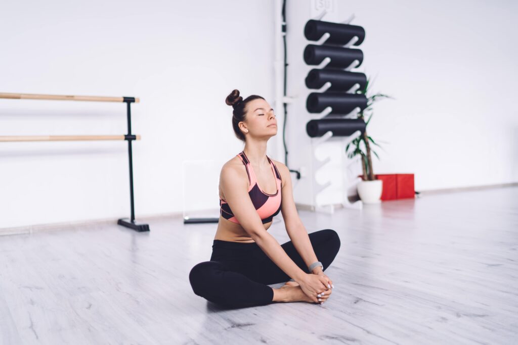 Calm relaxing fit female in sportswear with bun hairstyle sitting in butterfly position with eyes closed in bright spacious class Private suite for luxury residential treatment in California